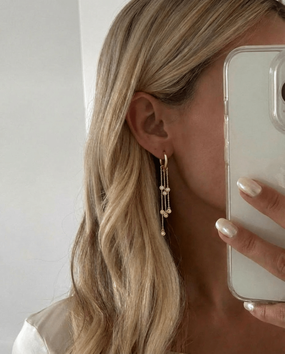 Mono boucle Golden Hour — créole chaînes asymétriques zircons bezel en argent 925 plaqué or 18k. 55mm, hypoallergénique, résistante à l'eau. Effet signature. vue portée par une femme blonde en selfie